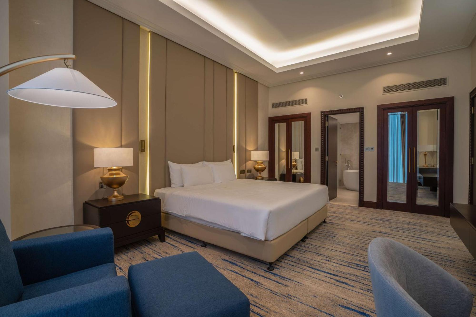 The Diplomat Radisson Blu Hotel Residence & Spa Manamah Buitenkant foto The image shows a modern hotel room. It features a queen-sized bed with white bedding, positioned against a backdrop of stylish wall panels. There are two bedside tables with lamps, and a large window is present, although it may be covered by curtain