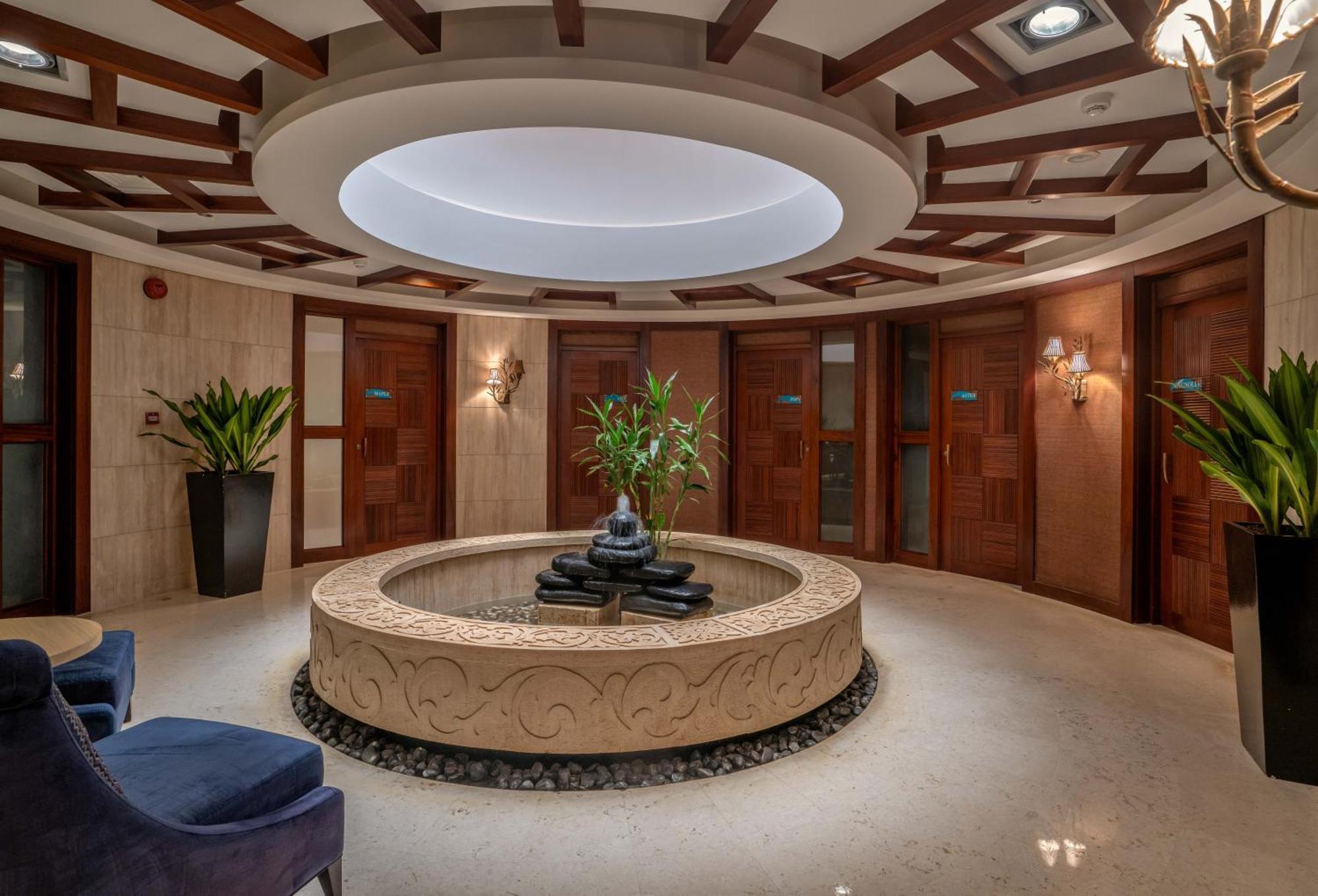 The Diplomat Radisson Blu Hotel Residence & Spa Manamah Buitenkant foto The photo shows a luxurious interior space, likely a lobby or relaxation area. The focal point is a circular stone basin containing a Buddha statue and smooth black stones, surrounded by a small bed of pebbles. The area is well-lit with a modern circ